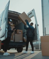 Workers loading boxes into a delivery van at a warehouse facility outdoors.